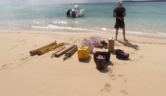 Le géomètre Antoine Germain se tenant fièrement sur une plage devant son matériel technique que vient de débarquer le bateau visible à l'arrière-plan