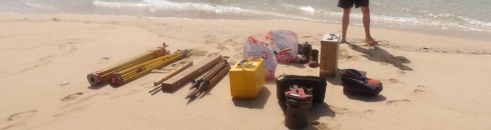Le géomètre Antoine Germain se tenant fièrement sur une plage devant son matériel technique que vient de débarquer le bateau visible à l'arrière-plan